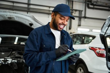 Mechanic checks vehicle details while smiling in a well equipped garage during busy daytime hours clipart