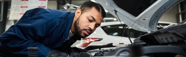 Odaklanmış genç bir tamirci, canlı bir oto tamir atölyesinde bir arabanın motorunu kontrol eder..