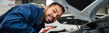 Genç bir tamirci canlı bir atölyede kaputun altındaki motoru incelerken gülümsüyor