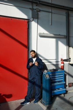 Şık, genç bir tamirci duvarın yanında duruyor, çalışırken telefonuna takılmış..