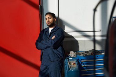 Handsome young mechanic leans against a wall, enjoying a moment of relaxation at work. clipart