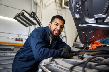 A skilled young mechanic smiles while inspecting an engine under a bright garage light clipart