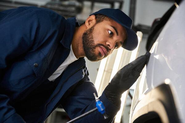 Красивый молодой механик тщательно осматривает автомобильную часть в ярком, хорошо оборудованном гараже