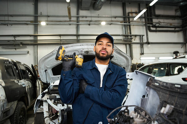Young man in blue work uniform stands proudly with tools in a bustling mechanic workshop.