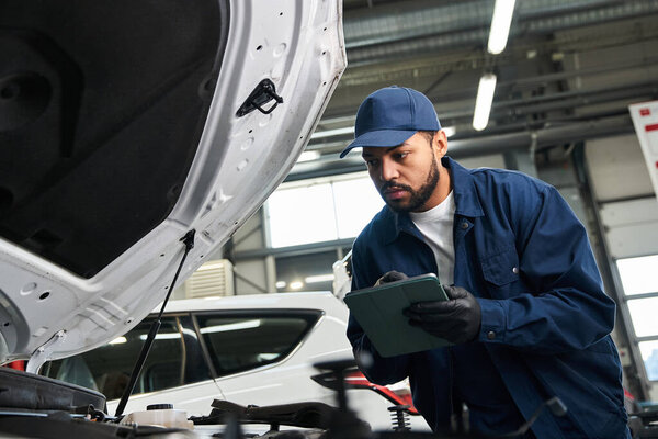 Сфокусированный молодой механик осматривает автомобильный двигатель, делая заметки в переполненной мастерской по ремонту автомобилей..