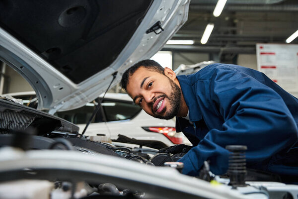 Молодой механик улыбается, мастерски чиня автомобильный двигатель под яркими лампами мастерской.