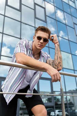 Handsome young man with tattoos and prosthetic leg stands on balcony against city backdrop. clipart