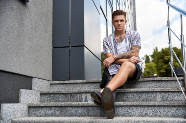 Handsome man with tattoos relaxes on outdoor stairs in an urban setting while enjoying the day. clipart