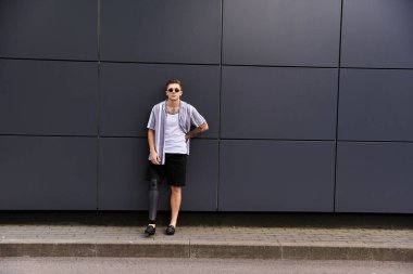 Young man with a prosthetic leg poses stylishly in front of a contemporary gray wall. clipart