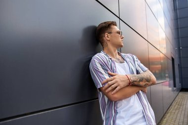 Dövmeleri olan yakışıklı genç adam, gösterişli bir duvara yaslanıyor..