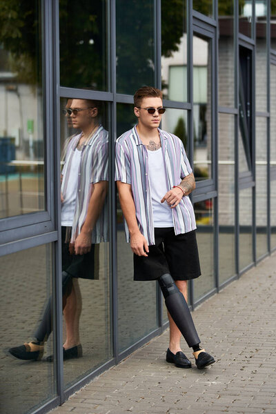 A young man with a prosthetic leg leans against glass windows, showcasing confidence and style.
