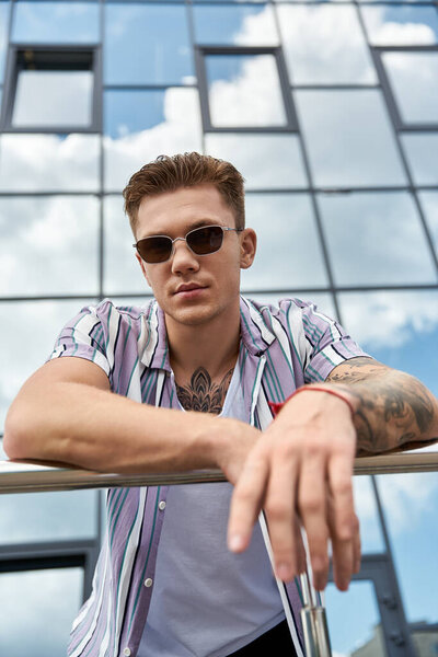 Расслабленный молодой человек демонстрирует свой стиль в очках и татуировках на солнце.