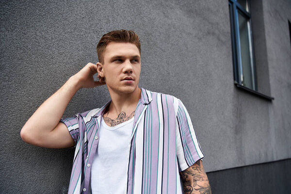 Молодой человек с татуировками уверенно позирует рядом с текстурированной серой стеной.