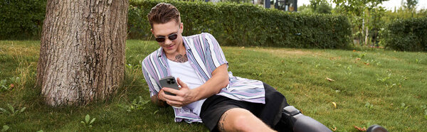Young man in sunglasses lies on grass, using his phone and enjoying a sunny day outdoors.