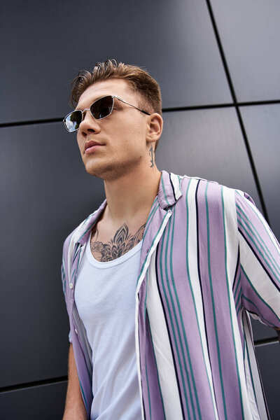 Молодой человек с татуировками уверенно позирует в модном наряде на современном фоне.