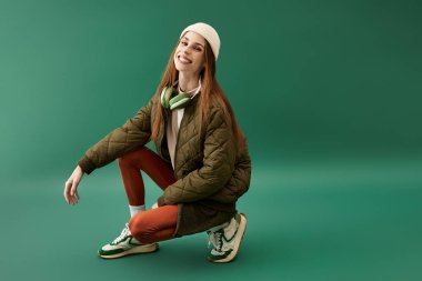 Parlak bir gülüşü olan genç bir kadın, yeşil bir arkaplanda şık bir şekilde poz veriyor..