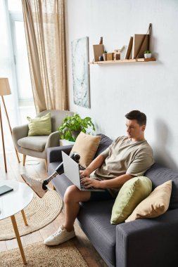 Genç adam evdeki işine odaklanmış, koltukta otururken dizüstü bilgisayar kullanıyor..