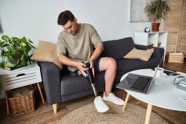 Handsome young man with prosthetic leg sits comfortably on a couch, focused on adjustment.