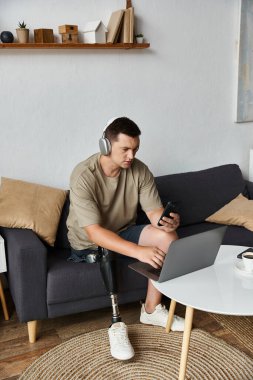 Protez bacaklı genç yakışıklı adam oturma odasında dizüstü bilgisayarla ve akıllı telefonla meşgul oluyor..