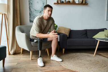 Handsome young man with a prosthetic leg sits thoughtfully in a modern living room. clipart