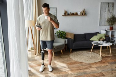 Yakışıklı genç adam şık oturma odasında duruyor, kahvesini yudumluyor ve akıllı telefonu kontrol ediyor..