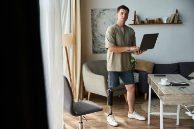 Oturma odasında dizüstü bilgisayar kullanırken protez bacaklı yakışıklı bir genç..