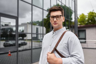 Handsome young man with stylish glasses stands outside a sleek building. clipart