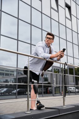 Protez bacaklı yakışıklı genç adam modern bir binanın dışında akıllı telefonunu kontrol ediyor..