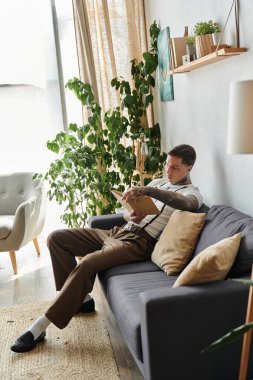 Dövmeleri olan yakışıklı genç bir adam rahat bir şekilde kanepede oturur ve kapalı alanda kitap okur..