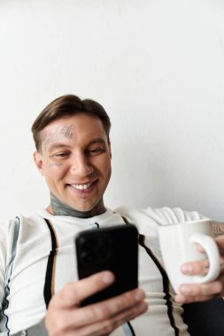 Man with tattoos smiles while sitting comfortably and checking his phone with a coffee mug. clipart