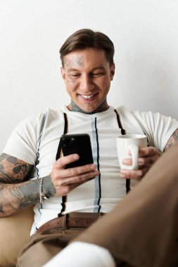 A young handsome man with tattoos smiles at his smartphone while holding a coffee cup. clipart