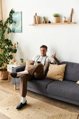 Tattooed young man relaxes on a couch, using his mobile in a stylish home. clipart