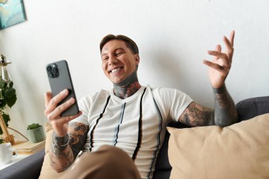 Dövmeli genç adam evde akıllı telefonunu kullanırken neşe içinde gülümsüyor..