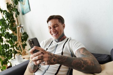 Dövmeli yakışıklı genç adam akıllı telefonunu açık ve rahat bir yaşam alanında kullanmaktan hoşlanıyor..