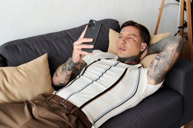 Dövmeli bir adam rahat bir odada akıllı telefonunu kontrol ederken kanepede öylesine uzanır..