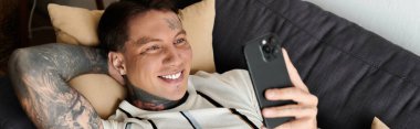 Relaxing young man with tattoos smiles as he looks at his phone on a comfortable couch. clipart