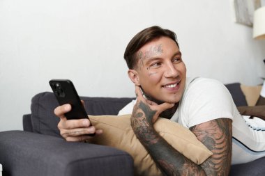 Yakışıklı bir adam, dövmeli bir kanepede uzanıyor, akıllı telefonunu kontrol ediyor ve gülümsüyor..