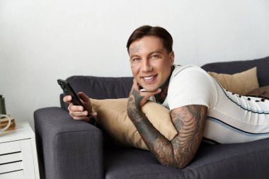 Dövmeli bir adam, akıllı telefonunu kanepede kullanırken rahatlamaktan zevk alır..
