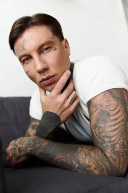 Yakışıklı genç bir adam, modern bir odada detaylı kol dövmeleri göstererek çenesini elinde dinlendiriyor..