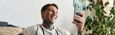 Dövmeli genç adam oturma odasında akıllı telefonunu kullanırken gülüyor..