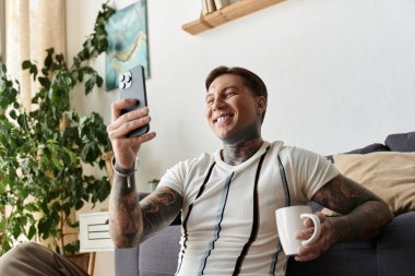 Dövmeleri olan mutlu genç adam koltukta dinleniyor, elinde kahve fincanı tutarken telefonunu kullanıyor..