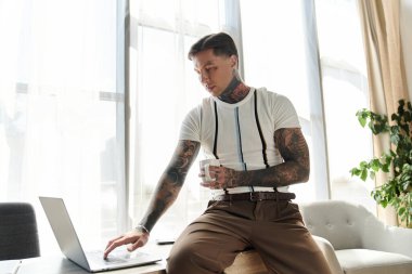Dövmeli genç bir adam şık bir odada elinde bardakla bir dizüstü bilgisayar kullanarak masada oturuyor..