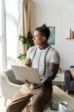 Dövmeli genç ve yakışıklı bir adam şık bir odada dizüstü bilgisayarına odaklanmış..