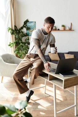 Handsome young man with tattoos sits at a desk using a laptop and sipping coffee in a cozy room. clipart