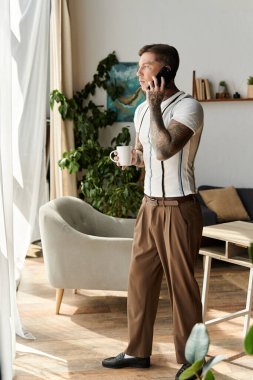 Handsome young man with tattoos stands by the window, talking on his phone and sipping coffee. clipart