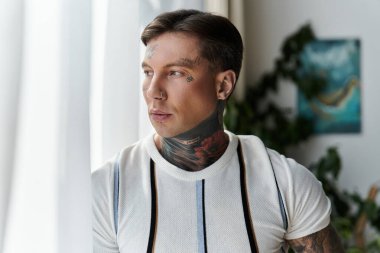 Dövmeleri olan yakışıklı genç adam iyi aydınlatılmış bir yerdeki pencereden dalgın dalgın bakıyor..