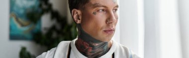 Young man with tattoos gazes thoughtfully while standing near a window with natural light. clipart