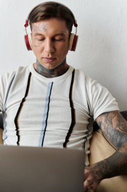 Dövmeleri olan genç bir adam evde dizüstü bilgisayarıyla çalışırken kulaklıkla müzik dinlemekten hoşlanır..