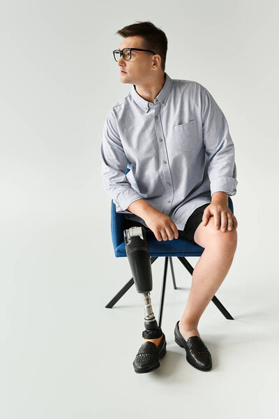 Young man with glasses and a prosthetic leg poses in a stylish chair in a simple studio.