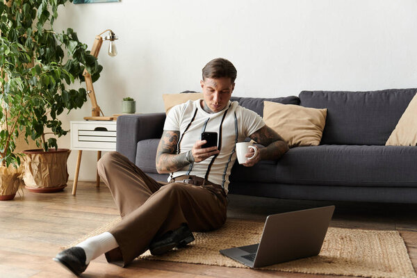 A young man with tattoos sits comfortably on the floor, holding a coffee cup and using his phone.